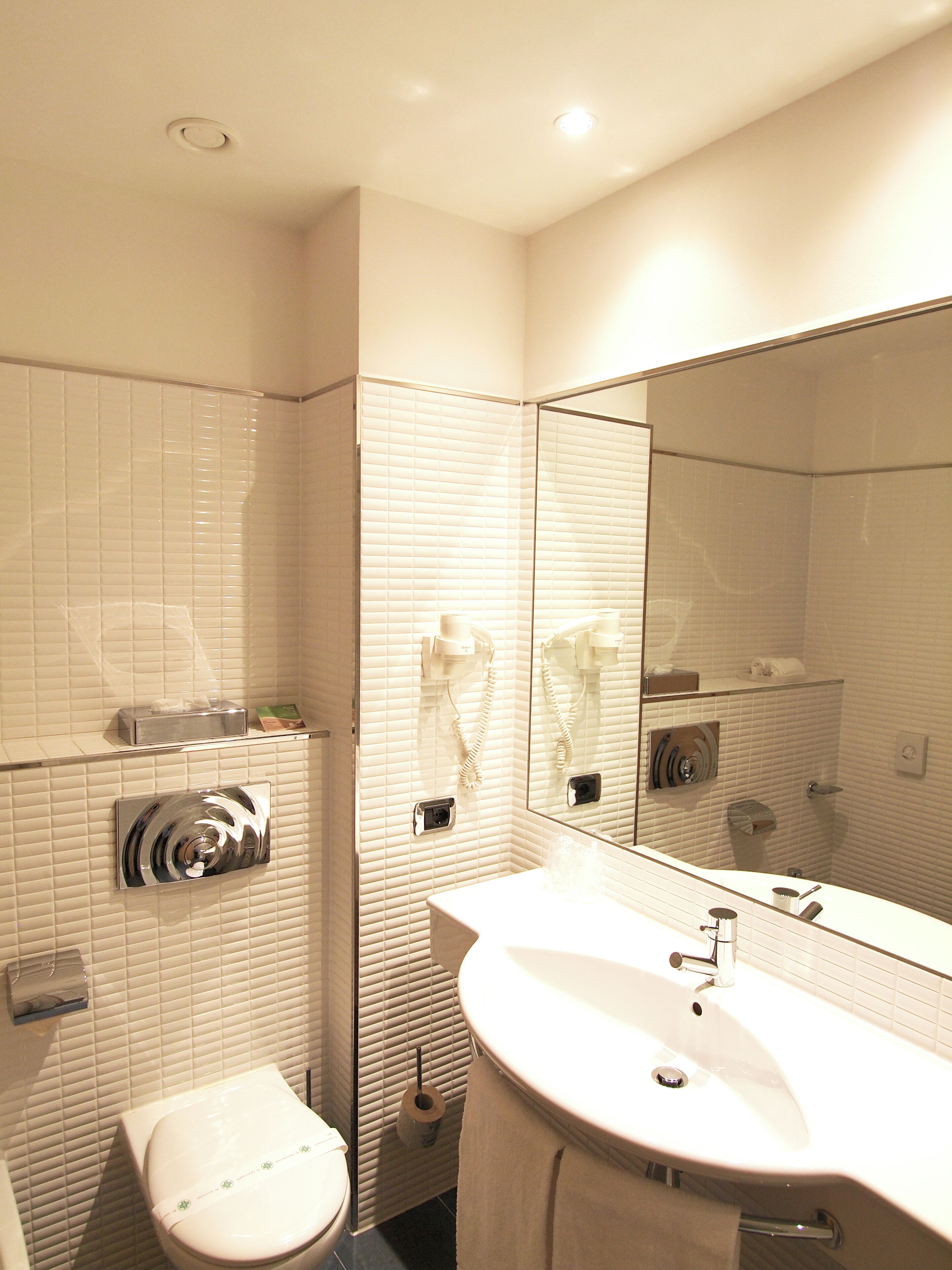 Ein modernes Badezimmer mit weißen Fliesen, einem großen Spiegel, einem Waschbecken und einer Toilette.
