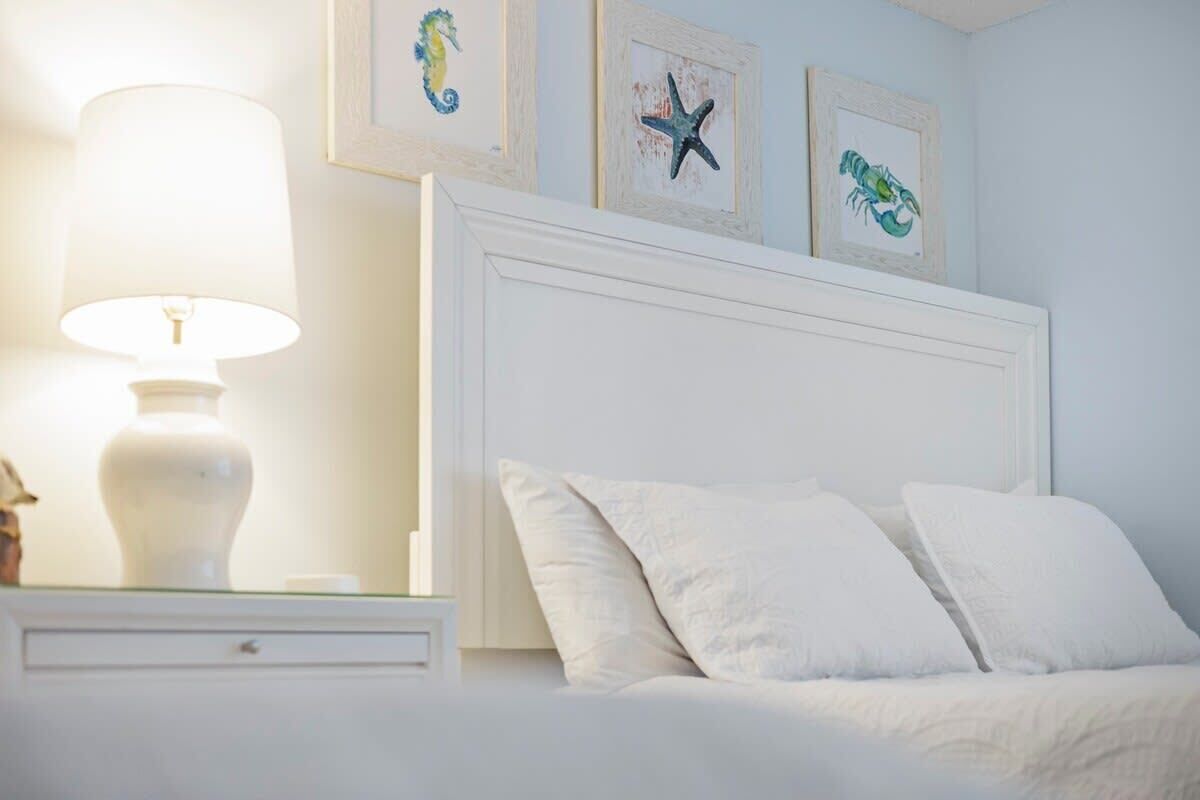 A neatly made bed with white bedding, a bedside lamp, and framed artwork on the wall.