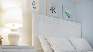 Ein ordentlich bezogenes Bett mit weißer Bettwäsche, eine Nachttischlampe und gerahmte Kunstwerke an der Wand.