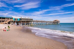 A beach with a pier extending into the water, featuring a restaurant and a shaded area.