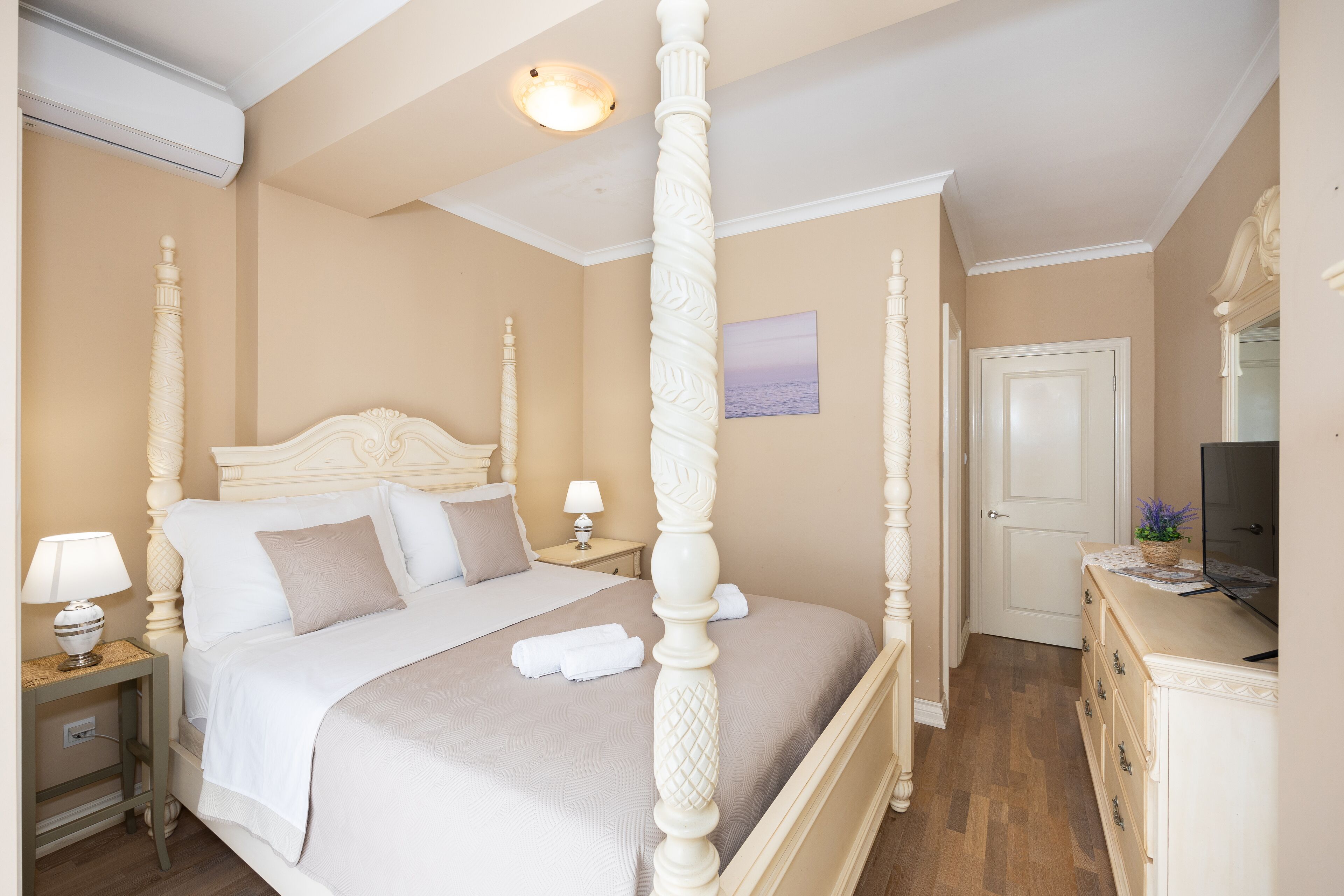 A four-poster bed with a headboard, a bedside table with a lamp, and a dresser with a flat-screen TV.