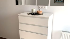 A white chest of drawers with a small table on top, a radiator, and three framed pictures on the wall. - Lovely 1 Bedroom Apartment (Kastela)
