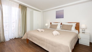 A neatly made bed with a beige bedspread and pillows, a wooden headboard, a nightstand with a lamp, and a window with sheer curtains. - Lovely 1 Bedroom Apartment (Kastela)