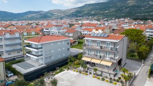 A multi-story hotel with balconies, a parking area, and a view of a residential area with red-roofed houses and mountains in the background. - Lovely 1 Bedroom Apartment (Kastela)