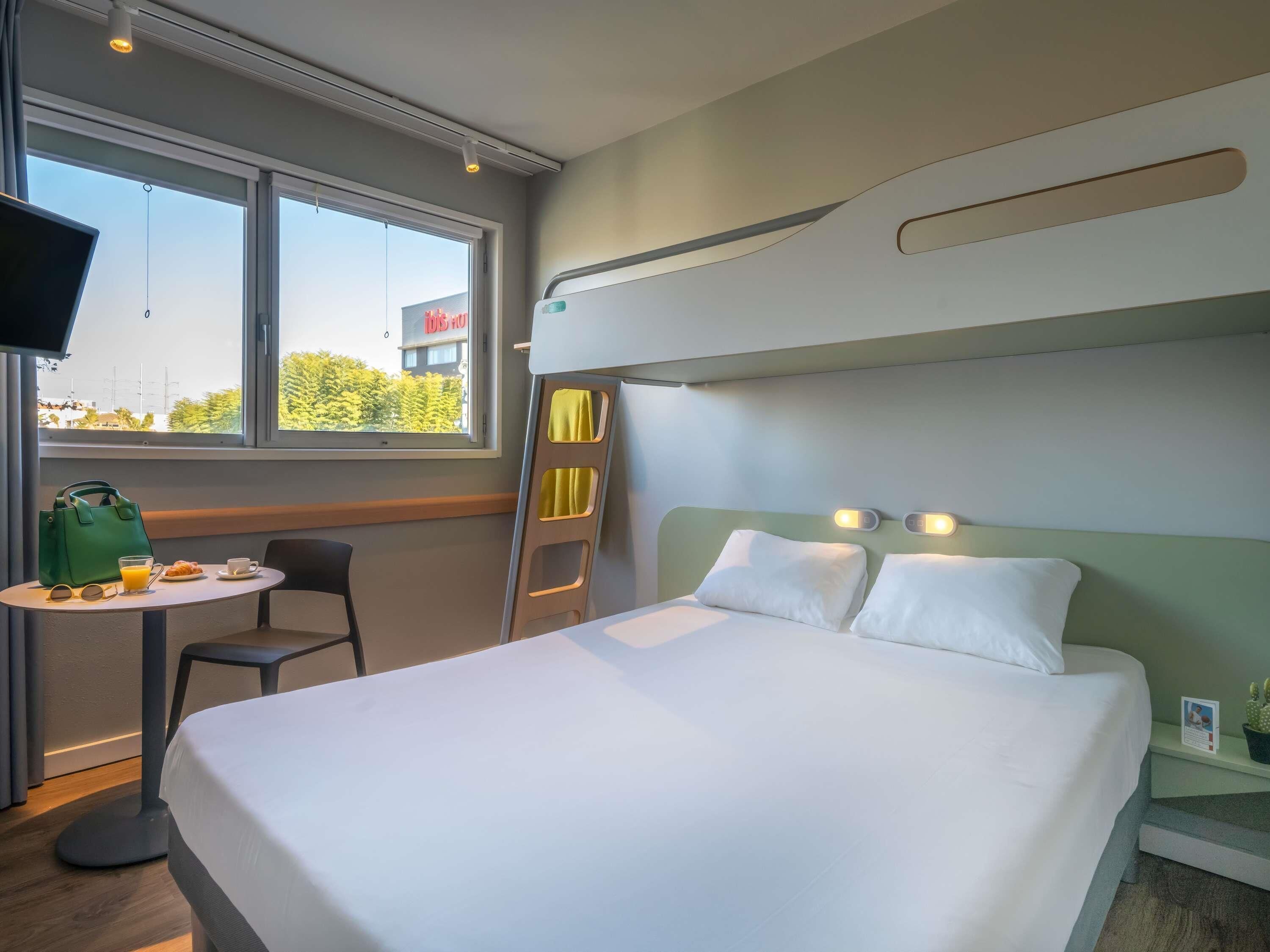 Une chambre d’hôtel avec un lit superposé, une petite table avec le petit-déjeuner, une chaise, une télévision et une fenêtre donnant sur des immeubles et des arbres.