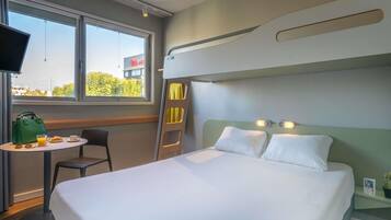Une chambre d’hôtel avec un lit superposé, une petite table avec le petit-déjeuner, une chaise, une télévision et une fenêtre donnant sur des immeubles et des arbres.