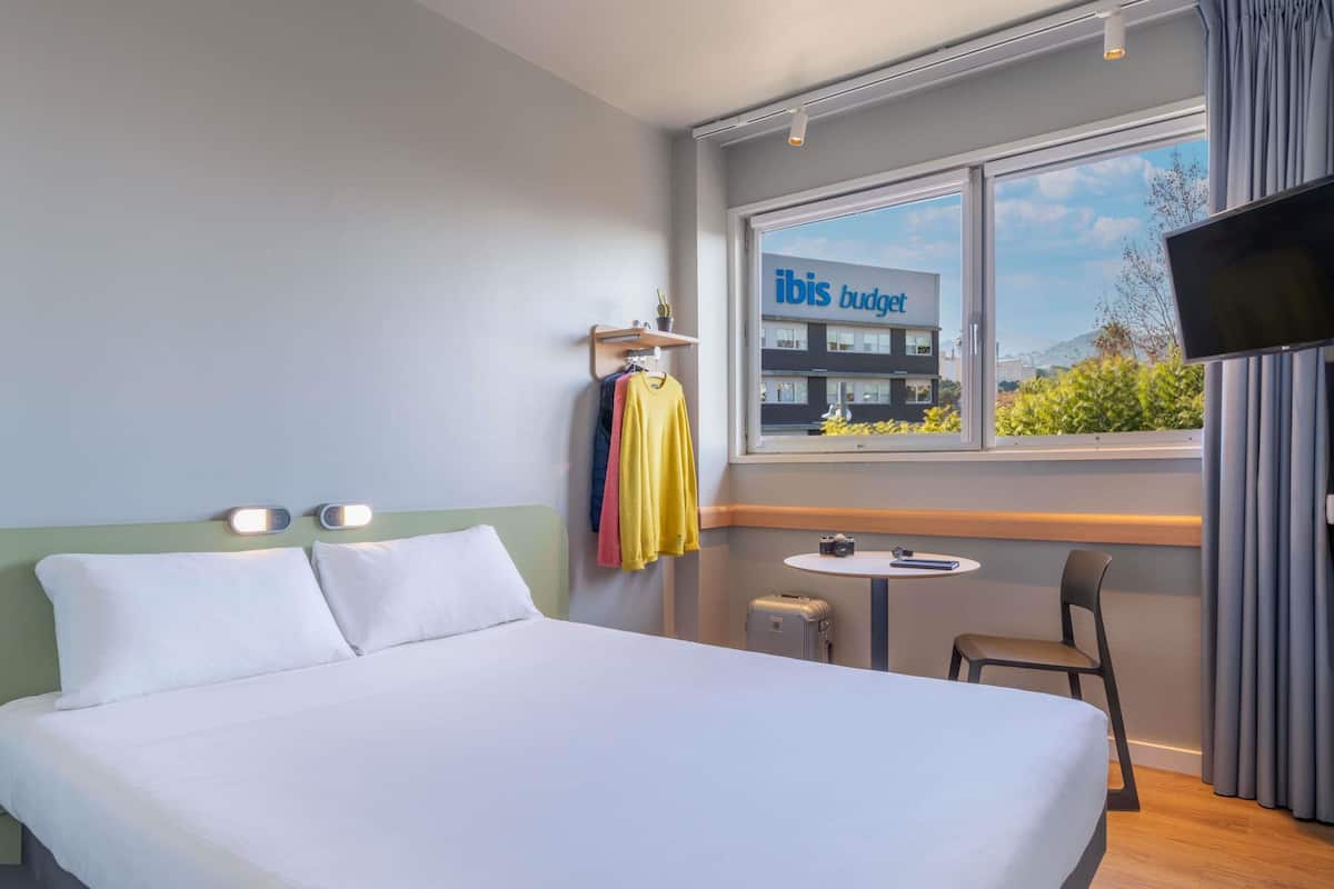 Une chambre d’hôtel avec un lit, une petite table sur laquelle se trouve un appareil photo, une chaise et une vue sur l’extérieur.