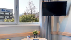 A hotel room with a bed, a small table with a plant, a TV, and a window with a view of a building and trees. - ibis Budget Aeropuerto Barcelona Viladecans (Viladecans)