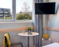 Quarto de hotel com cama, uma pequena mesa com uma planta, televisão e janela com vista para um edifício e árvores.