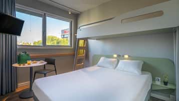 Habitación de hotel con cama, una mesita con desayuno, una silla, televisor y ventana con vistas a edificios y árboles.