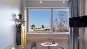 A hotel room with a bed, a small table with a chair, a TV, a window with a view of palm trees and buildings, and a suitcase. - ibis Budget Aeropuerto Barcelona Viladecans (Viladecans)