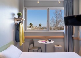 Quarto de hotel com cama, mesa pequena com cadeira, televisão, janela com vista para palmeiras e edifícios, e uma mala.