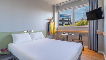 Une chambre d’hôtel avec un lit, une petite table, une chaise, un téléviseur et une fenêtre donnant sur un bâtiment portant l’inscription « ibis budget ».