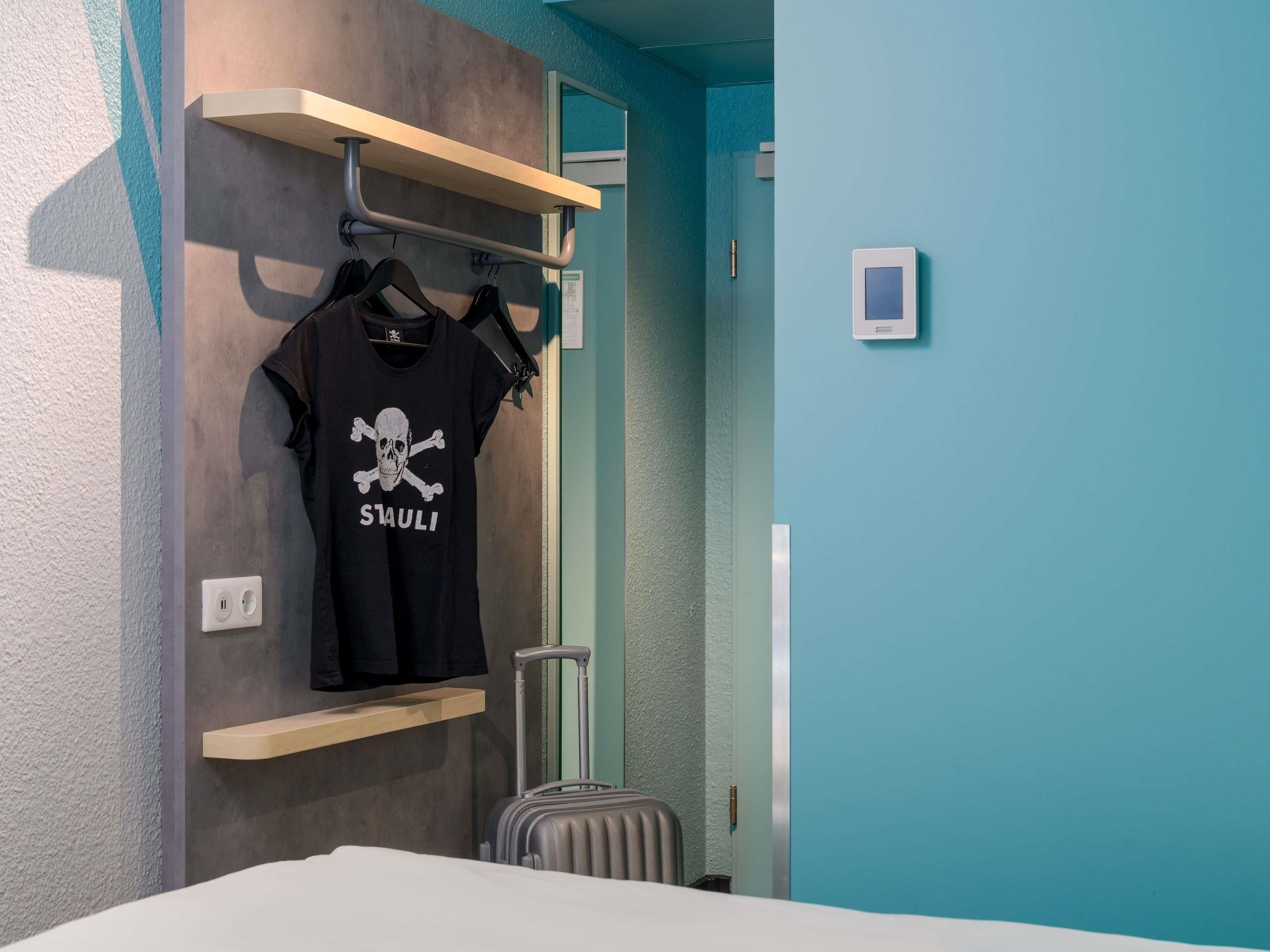 Ein Hotelzimmer mit einem Bett, einem Koffer, einem an der Wand montierten Regal mit einem schwarzen T-Shirt und einer Tür mit Griff.