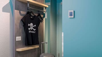 Ein Hotelzimmer mit einem Bett, einem Koffer, einem an der Wand montierten Regal mit einem schwarzen T-Shirt und einer Tür mit Griff.