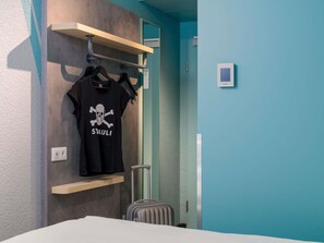 A hotel room with a bed, a suitcase, a wall-mounted shelf with a black t-shirt, and a door with a handle. - Ibis Budget Hamburg St Pauli Messe (Hamburg)
