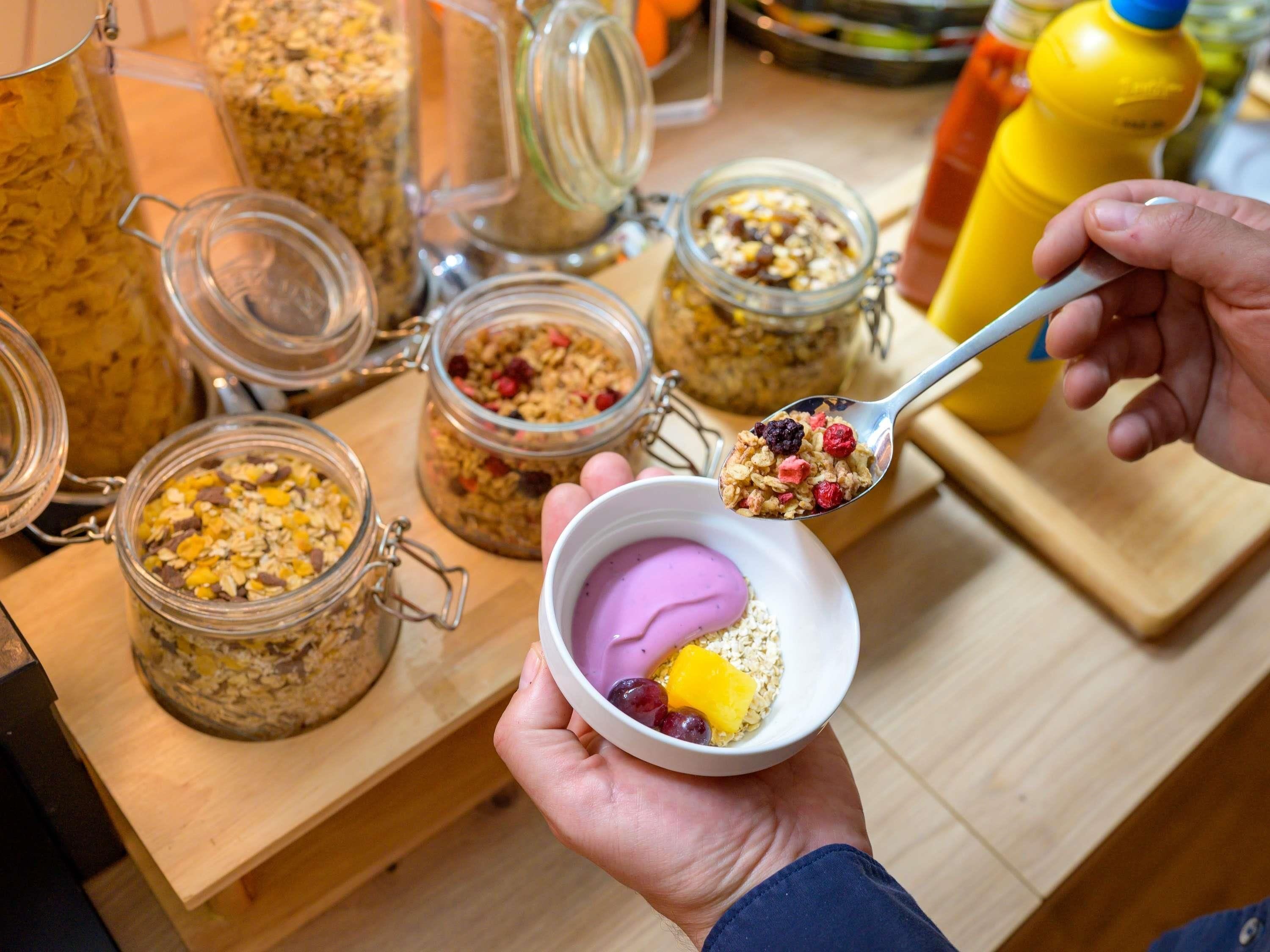Una persona sosteniendo un tazón con una cucharada de granola y yogur, con botes de granola al fondo.