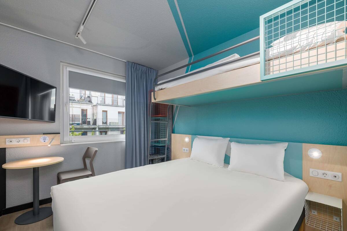 Una cama litera con un televisor encima, una mesita pequeña y una ventana con cortinas.