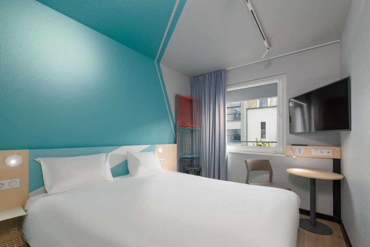 Habitación de hotel con una cama grande, un televisor en la pared, una mesita y una ventana con cortinas.