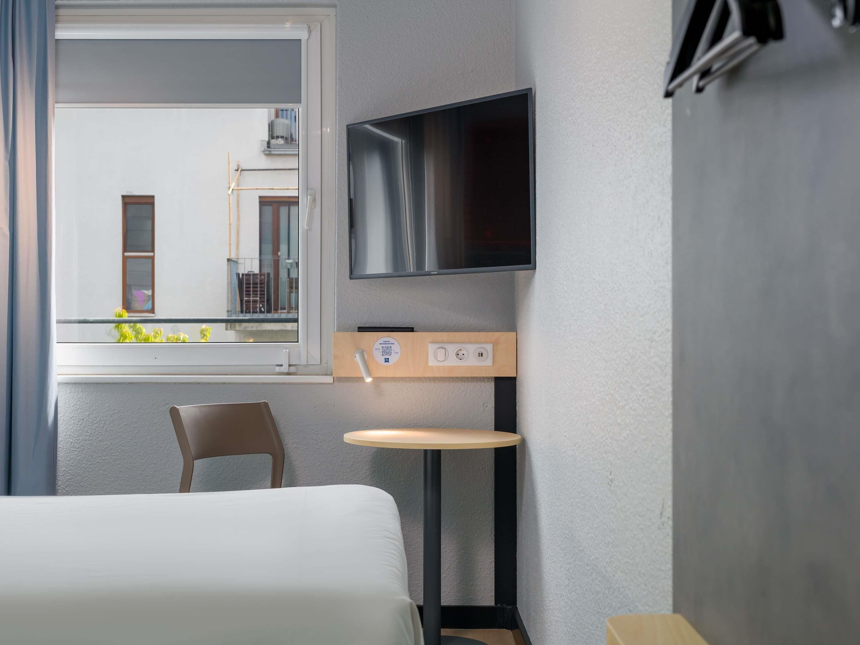 Ein Hotelzimmer mit einem Bett, einem Sessel, einem kleinen Tisch, einem an der Wand befestigten Fernseher und einem Fenster mit Blick ins Freie.