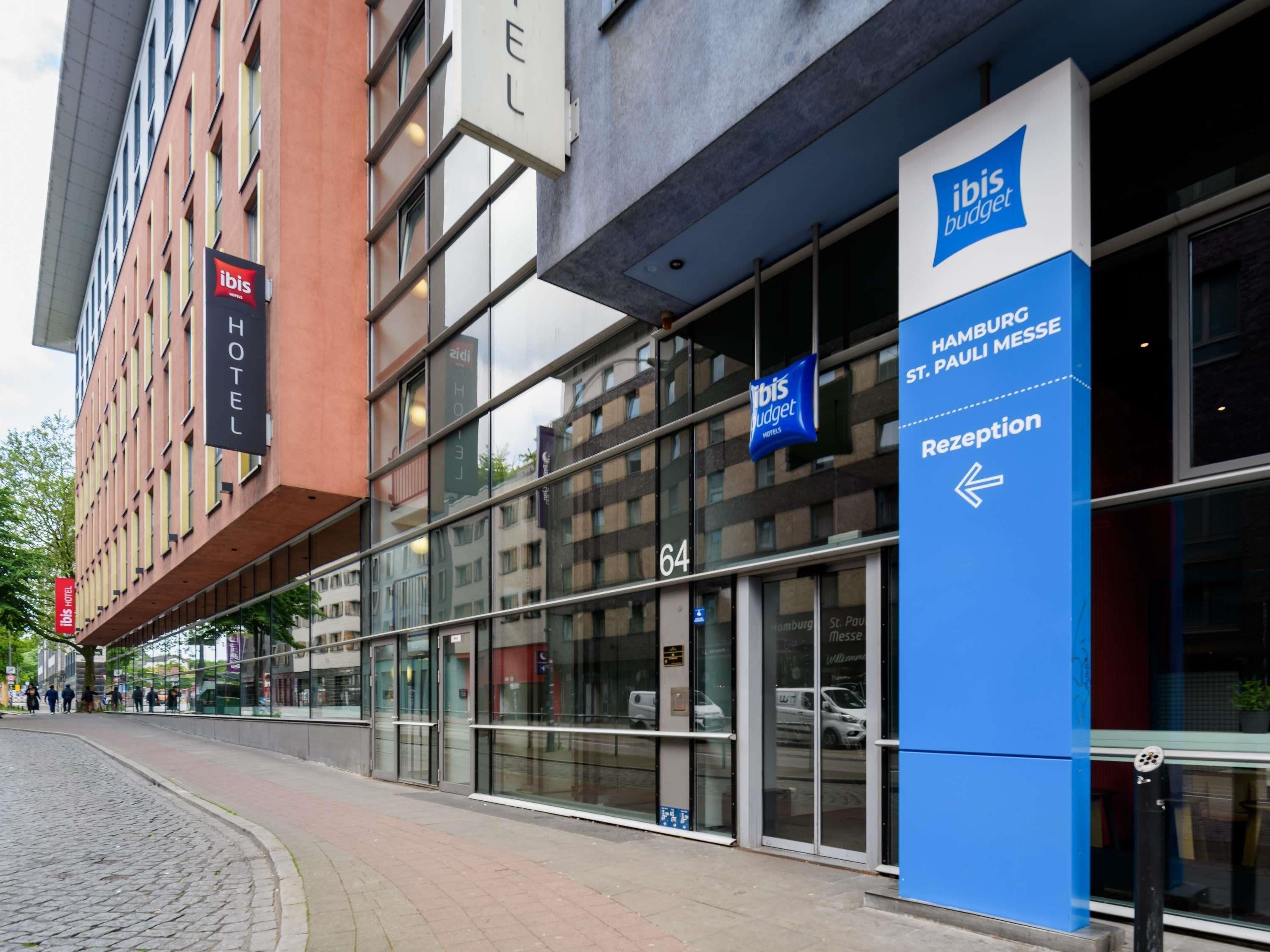 A modern building with a glass facade, a sign for 'Ibis Hotel,' and a blue sign indicating 'Reception' for 'HAMBURGO St. PAULI MESSE.'