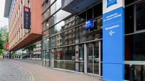 Ein modernes Gebäude mit Glasfassade, einem Schild mit der Aufschrift 'Ibis Hotel' und einem blauen Schild, das 'Rezeption' für 'HAMBURGO St. PAULI MESSE' anzeigt.