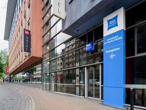 Een modern gebouw met een glazen gevel, een bord met 'Ibis Hotel' en een blauw bord waarop 'Receptie' voor 'HAMBURGO St. PAULI MESSE' staat.