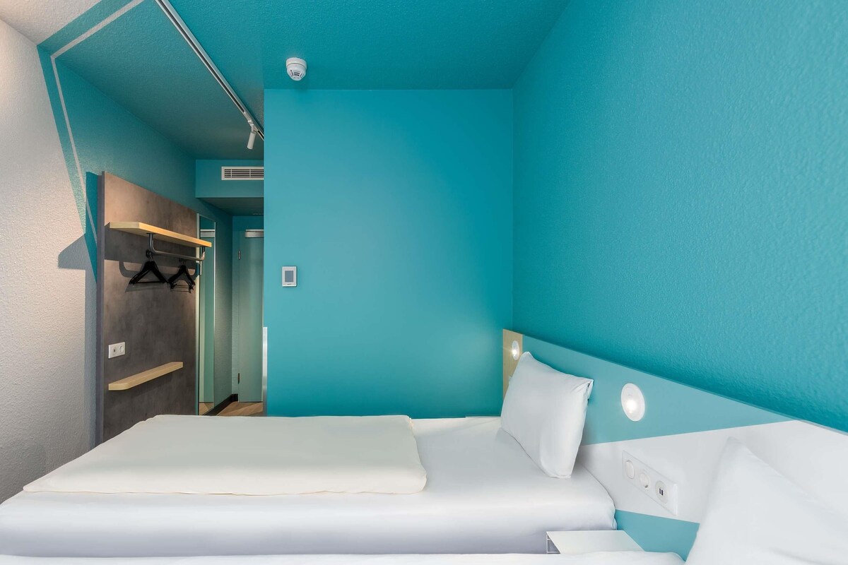Ein Zimmer mit einer türkisfarbenen Wand, einem Bett mit weißer Bettwäsche, einem Kleiderständer und einer Tür zu einem anderen Raum.