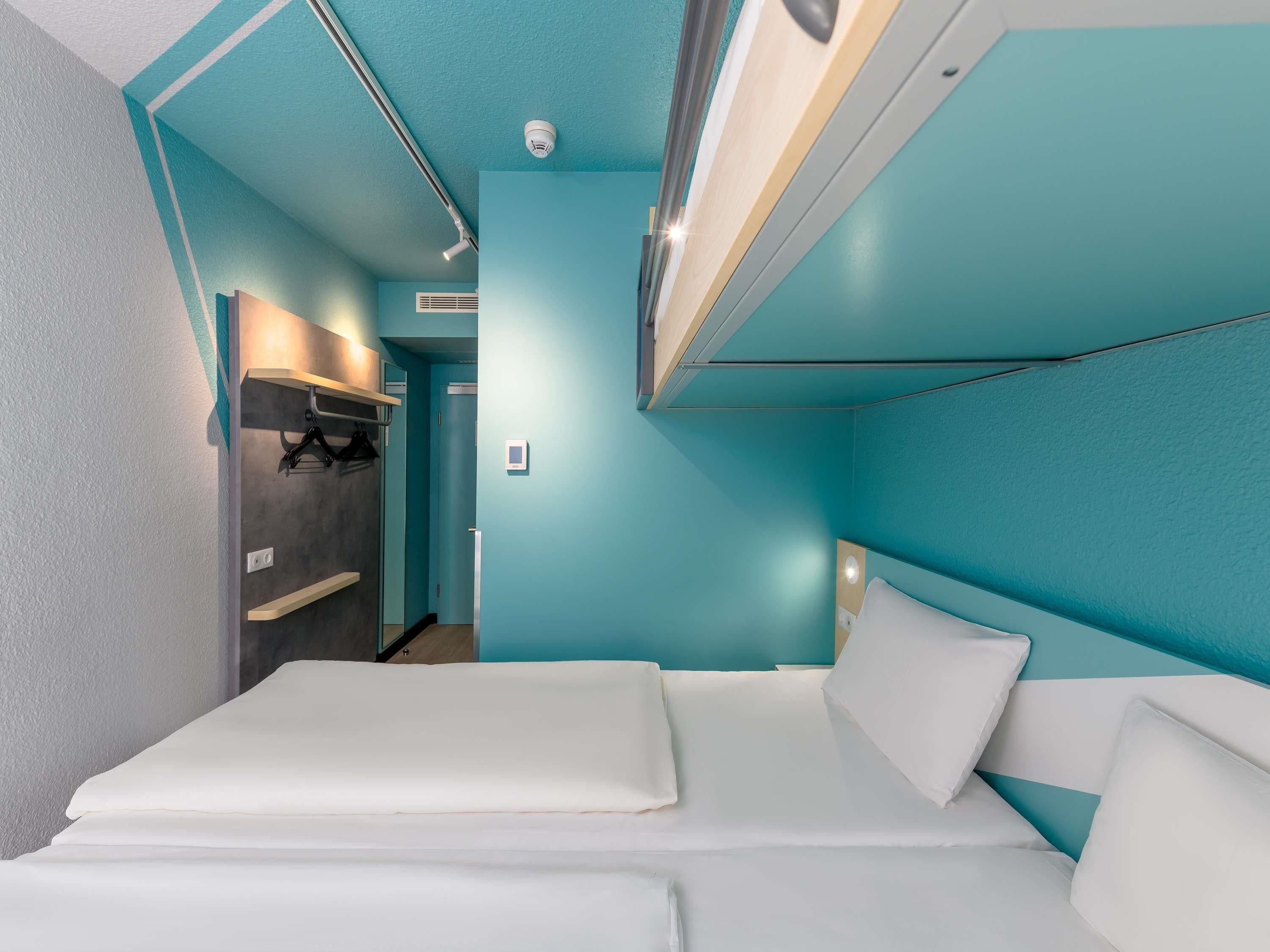 A modern hotel room with teal walls, a bed with white linens, a built-in wardrobe, and a ceiling-mounted light fixture.
