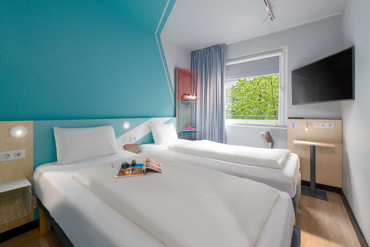 Ein Hotelzimmer mit Bett, Nachttisch, Fernseher, Fenster und Blick auf Grünfläche.