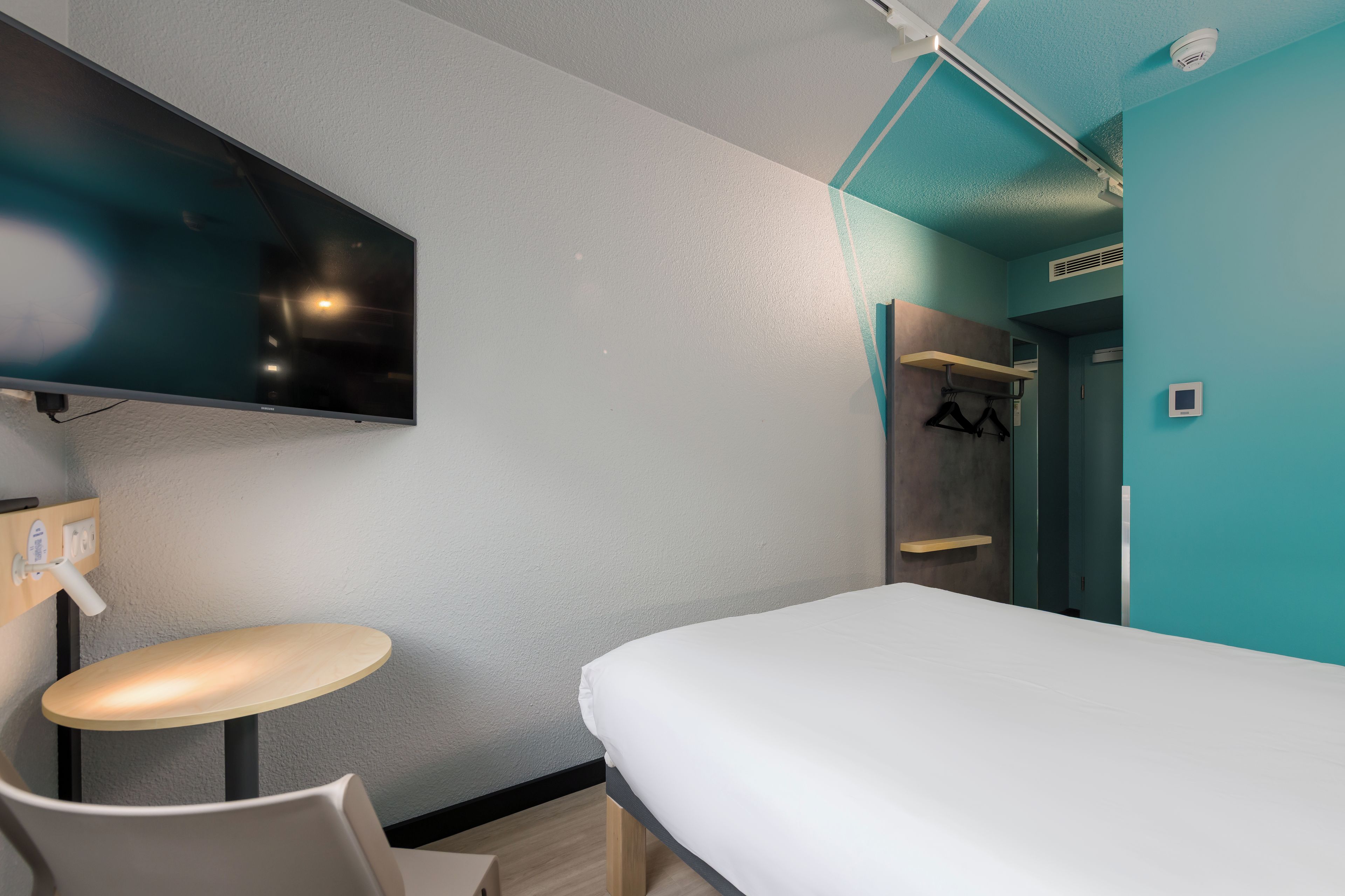 A hotel room with a bed, a TV mounted on the wall, a small round table, and a mirror.