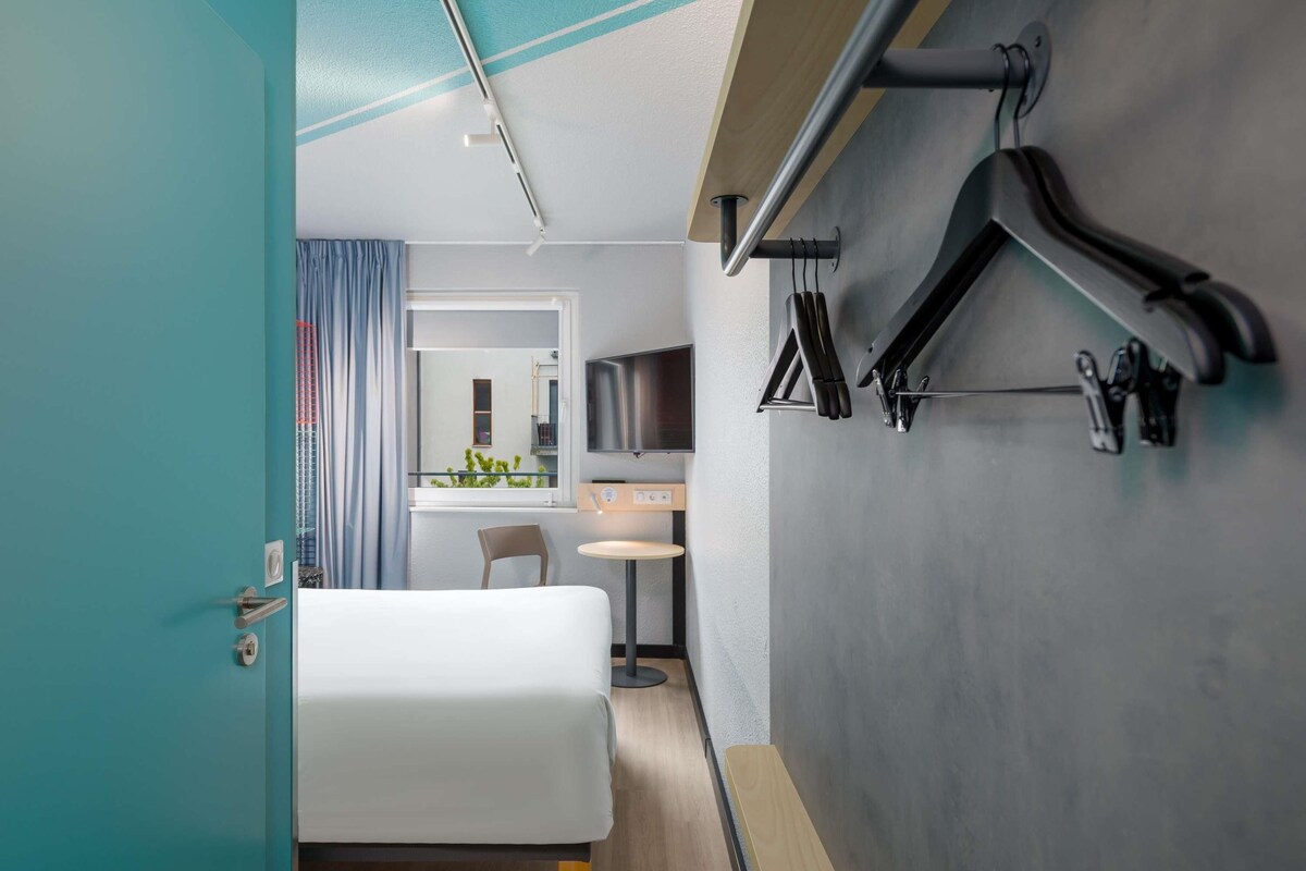 Ein Hotelzimmer mit Bett, Schreibtisch, Stuhl, Fernseher und einem an der Wand befestigten Ablagefach mit Gegenständen.