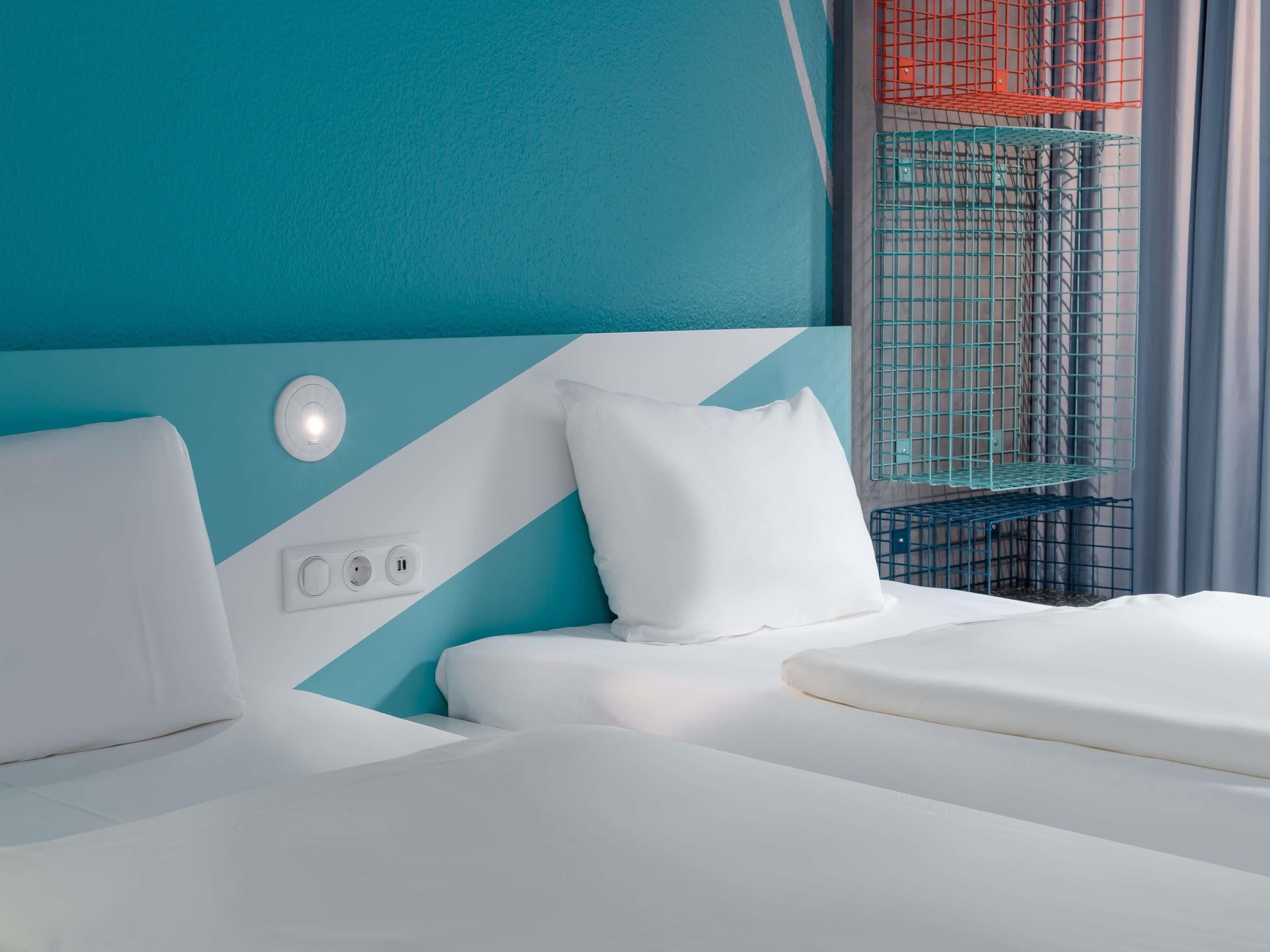 Habitación de hotel con cama, enchufe y ventana con celosía metálica.