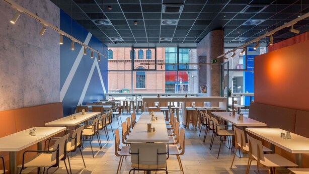 Un comedor amplio con varias mesas y sillas, ventanas grandes que ofrecen vistas a un edificio de ladrillo y un diseño de techo moderno.