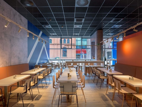 A spacious dining area with multiple tables and chairs, large windows offering views of a brick building, and a modern ceiling design.