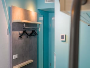 Een gang met een turquoise wand, een kapstok met hangers, een lichtschakelaar en een thermostaat aan de muur.