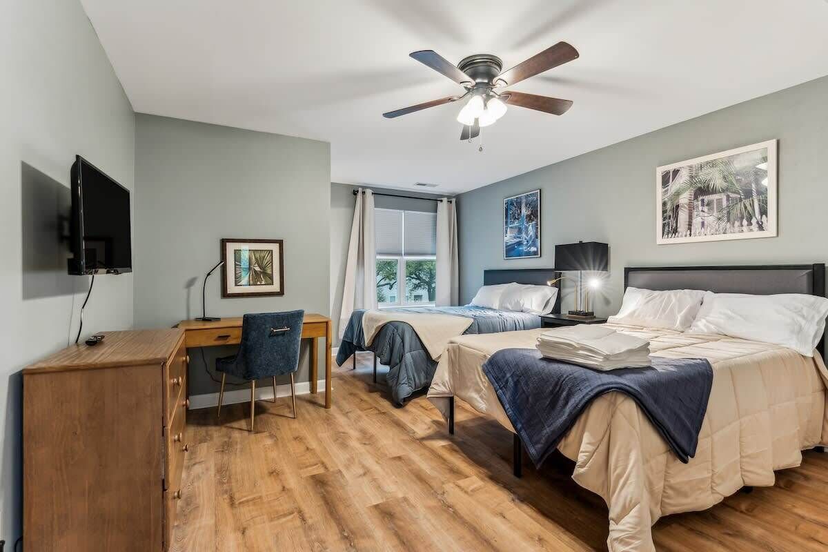 A bedroom with two beds, a desk, a chair, a ceiling fan, and a TV.
