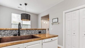 Una cocina con encimera de madera, un lavamanos y un lavavajillas. Hay una ventana con persianas, un colgante de luz y una puerta blanca.