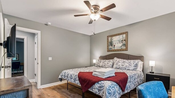 Un dormitorio con cama, ventilador de techo, mesita de noche, una silla y un cuadro en la pared.