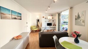 Ein modernes Wohnzimmer mit einer Couch, einem Esstisch und einem Fernseher. Ein Fenster bietet Blick ins Freie.