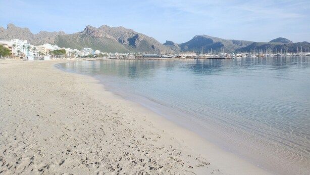 Ein Strand mit klarem Wasser, eine Marina mit Booten und Berge im Hintergrund.