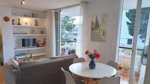 Ein modernes Wohnzimmer mit einem grauen Sofa, einem runden Esstisch mit weißen Stühlen, einem Fernseher auf einem Regal und einem Balkon mit Blick auf Gebäude und Bäume.