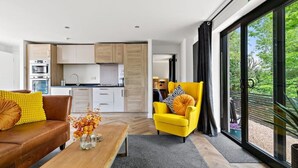 Una sala moderna con un sofá, un sillón amarillo y una mesita de centro. Se aprecia una cocina al fondo con armarios blancos y encimera negra.