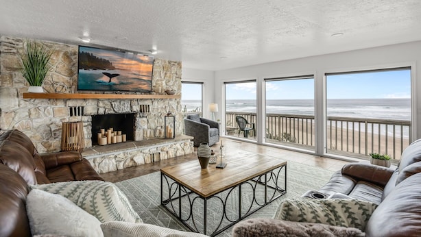 A spacious living room with a stone fireplace, leather sofas, a large TV, and a balcony with a view of the ocean. - Azul Jewel Estate, Game Room, Movie Room & Beach (Depoe Bay)
