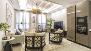 Una sala de estar moderna con un sofá, dos sillones de mimbre negro, una mesa de comedor, un televisor y una zona de cocina con nevera y horno.