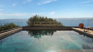 A clear view of a swimming pool with a glass bottom, surrounded by a wooden deck and a view of the sea and distant mountains.