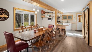 Un coin repas avec une table et des chaises en bois, une cuisine avec des meubles blancs et une vue sur l’extérieur grâce à de grandes fenêtres.