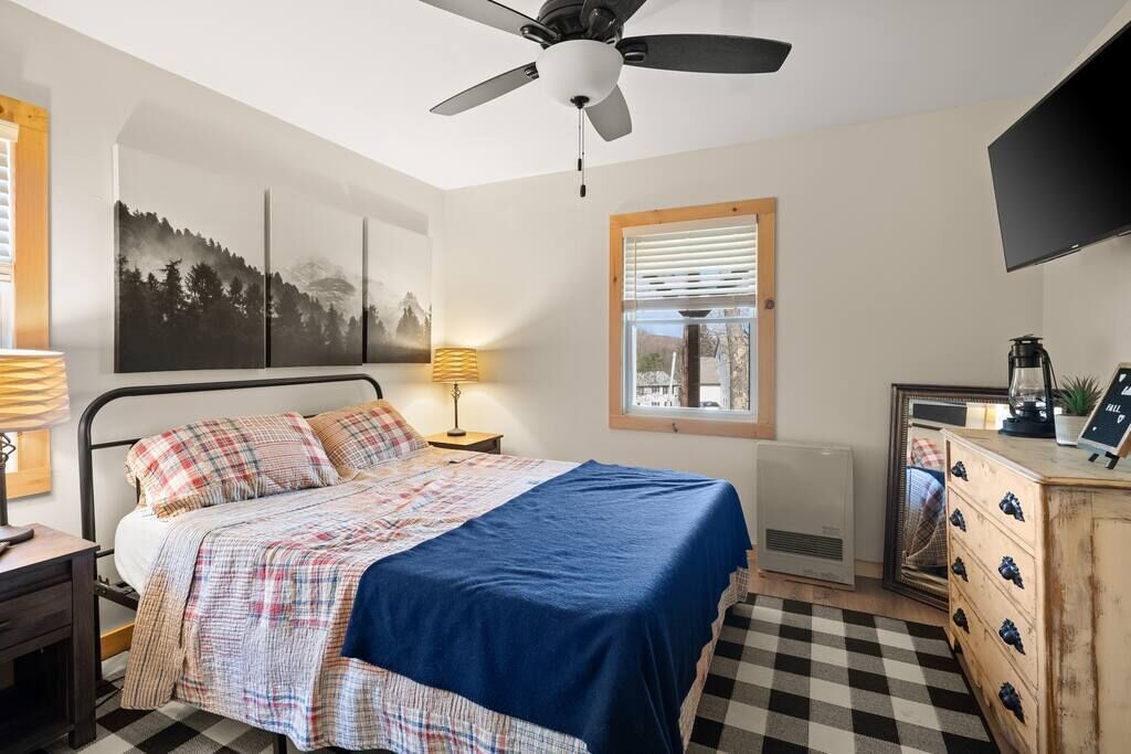 Ein Schlafzimmer mit Bett, Deckenventilator, Fenster mit Jalousien, Heizkörper, Kommode und einem an der Wand befestigten Fernseher.
