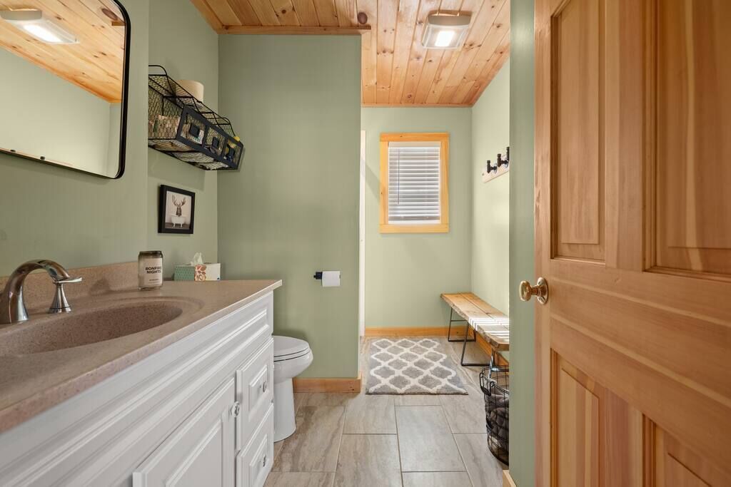 Une salle de bain avec un plafond en bois, un lavabo avec un miroir, des toilettes, une fenêtre avec des stores et un banc.