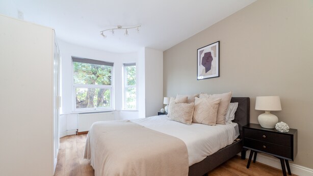 A bedroom with a large bed, a bedside table with a lamp, a framed picture on the wall, and a window with a view of trees.